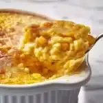 Spoonful of corn pudding being lifted from white ceramic dish showing layers of golden crust and creamy corn-studded interior on marble surface