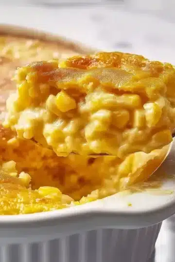 Spoonful of corn pudding being lifted from white ceramic dish showing layers of golden crust and creamy corn-studded interior on marble surface