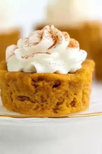 Macro close-up of keto pumpkin pie cupcake on white plate with gold rim, showing detailed cake texture and fluffy whipped cream topping dusted with pumpkin pie spice