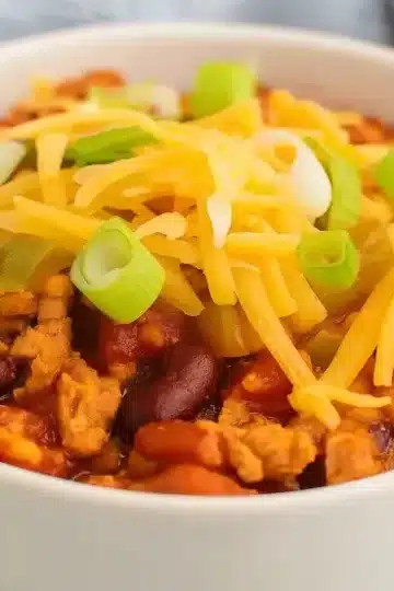 White bowl filled with turkey chili topped with melted cheddar cheese and fresh green onions, showing kidney beans and ground turkey in tomato sauce