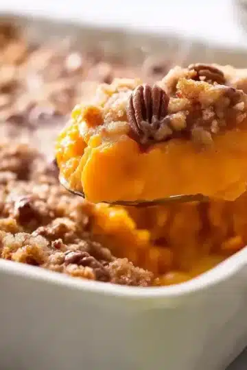 Diagonal view of silver spoon lifting portion of sweet potato casserole topped with whole pecan half from white ceramic baking dish