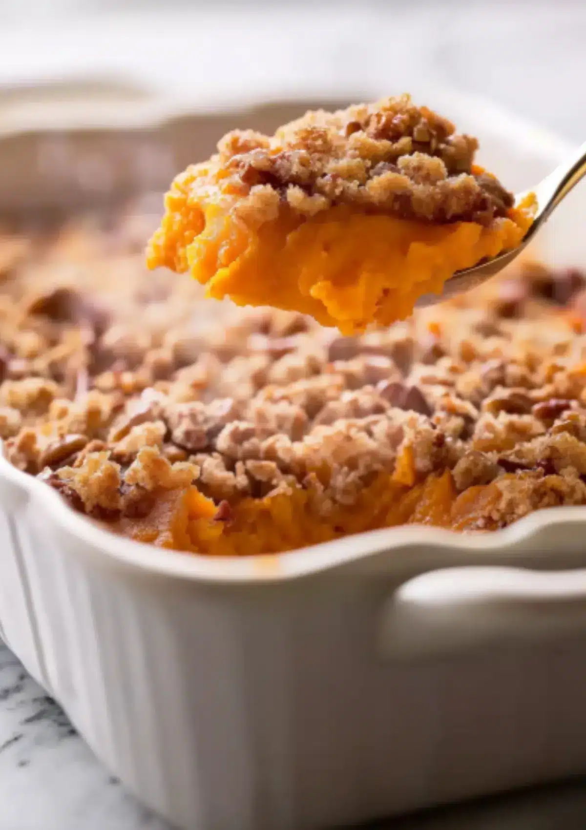 Spoonful of sweet potato casserole lifted from white ceramic baking dish revealing creamy orange sweet potato layer beneath golden brown pecan streusel topping