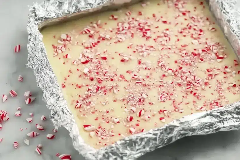 Overhead view of uncut white chocolate peppermint fudge in foil-lined baking pan topped with crushed candy canes ready to be sliced