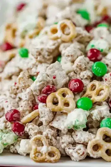 Reindeer chow with thick white chocolate coating on Cheerios, pretzels, peanuts and M&Ms served on white platter with pink textured napkin