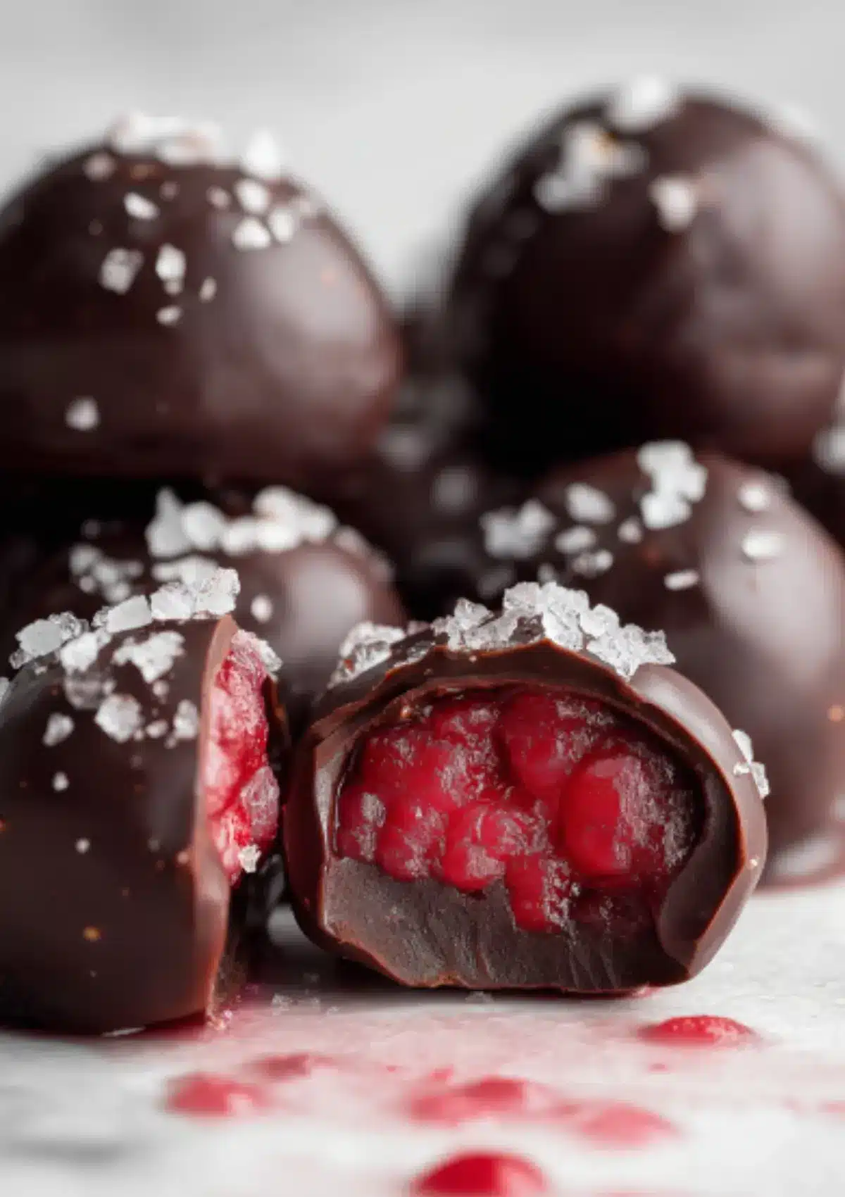 Macro close-up of chocolate covered raspberry chia bite broken open showing bright crimson red filling with visible chia seeds, topped with flaky sea salt, white marble background