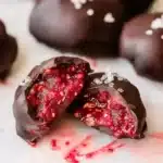 Close-up macro shot of chocolate covered raspberry chia bite cut in half showing textured bright red raspberry filling with visible chia seeds, glossy dark chocolate shell, white marble background with whole bites blurred in background