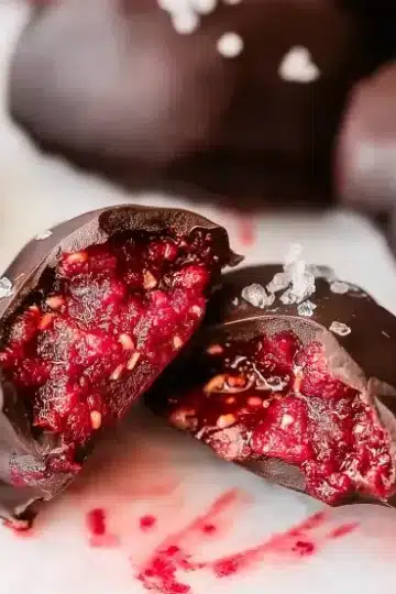 Close-up macro shot of chocolate covered raspberry chia bite cut in half showing textured bright red raspberry filling with visible chia seeds, glossy dark chocolate shell, white marble background with whole bites blurred in background