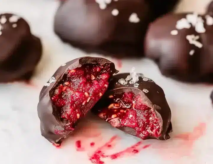 Close-up macro shot of chocolate covered raspberry chia bite cut in half showing textured bright red raspberry filling with visible chia seeds, glossy dark chocolate shell, white marble background with whole bites blurred in background
