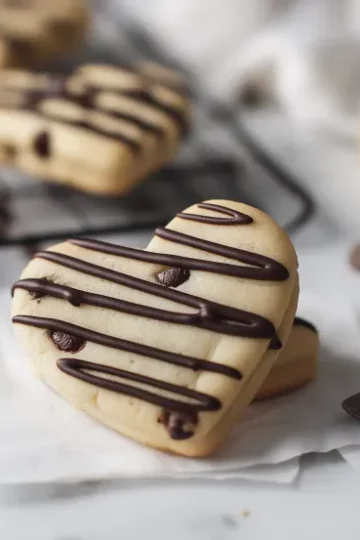 Overhead view of heart-shaped chocolate chip sugar cookies arranged on a wire cooling rack with white parchment paper, some decorated with dark chocolate drizzle stripes, mini chocolate chips scattered around