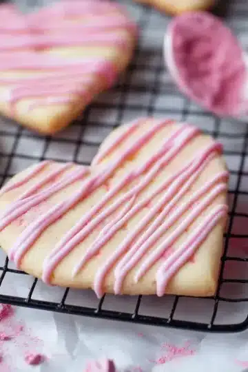 Macro close-up photograph of heart-shaped strawberry shortbread cookies on a black wire cooling rack with delicate pink and white chocolate drizzle patterns, surrounded by pink strawberry powder, measuring spoon, and heart-shaped cookie cutter on white marble surface with shallow depth of field
