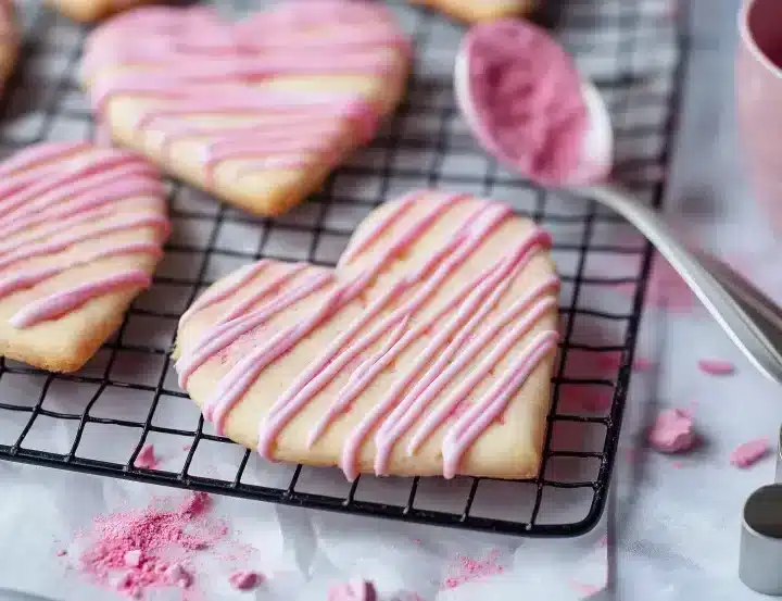 Macro close-up photograph of heart-shaped strawberry shortbread cookies on a black wire cooling rack with delicate pink and white chocolate drizzle patterns, surrounded by pink strawberry powder, measuring spoon, and heart-shaped cookie cutter on white marble surface with shallow depth of field