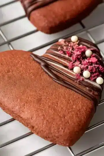 Heart-shaped chocolate Valentine cookies on wire cooling rack, decorated with elegant dark chocolate drizzle, pink freeze-dried raspberries, and white chocolate pearls