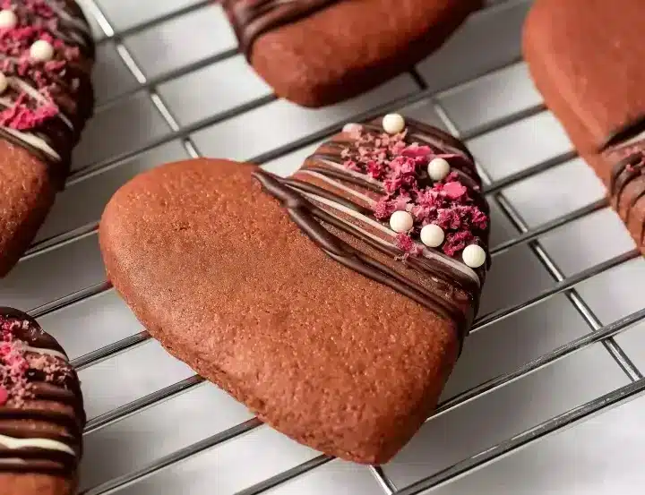 Heart-shaped chocolate Valentine cookies on wire cooling rack, decorated with elegant dark chocolate drizzle, pink freeze-dried raspberries, and white chocolate pearls