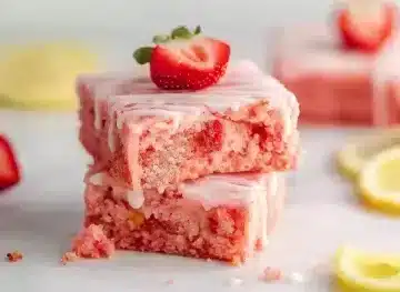 Two pink strawberry brownies stacked on white surface showing crumbly texture with white lemon glaze and fresh strawberry garnish, lemon slices in background