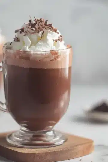 Rich homemade hot chocolate served in a clear glass mug with generous whipped cream topping and dark chocolate shavings, placed on a wooden coaster against a soft gray marble background
