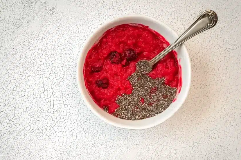 White ceramic bowl containing bright red mashed fresh raspberries with black chia seeds scattered on top, vintage silver spoon resting in bowl, overhead view on crackled marble surface