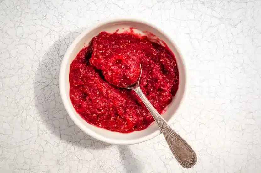 White ceramic bowl containing vibrant red raspberry chia jam with chia seeds fully incorporated throughout, vintage silver spoon in bowl, overhead view on textured crackled surface