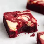 Close-up of a single red velvet cheesecake brownie square showing cream cheese swirl on top and fudgy dense interior texture, with additional brownies blurred in background on white marble surface