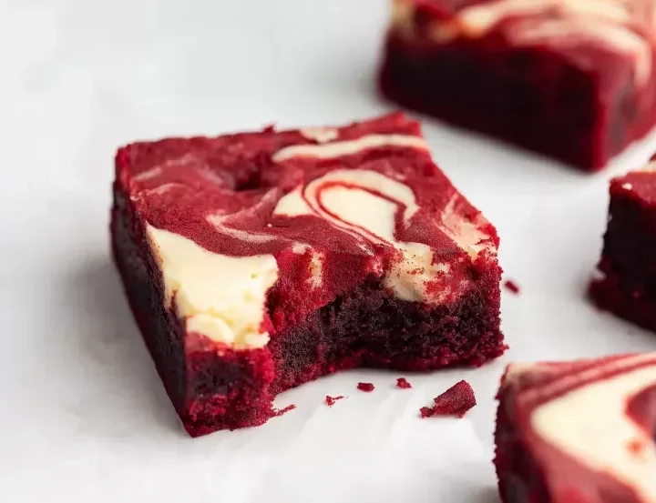 Close-up of a single red velvet cheesecake brownie square showing cream cheese swirl on top and fudgy dense interior texture, with additional brownies blurred in background on white marble surface