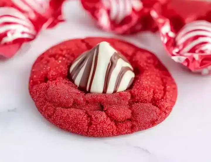 Close-up of one red velvet cookie with sparkly crackled texture topped with white and brown striped Hershey's Kiss, red and white candy wrappers softly blurred in background