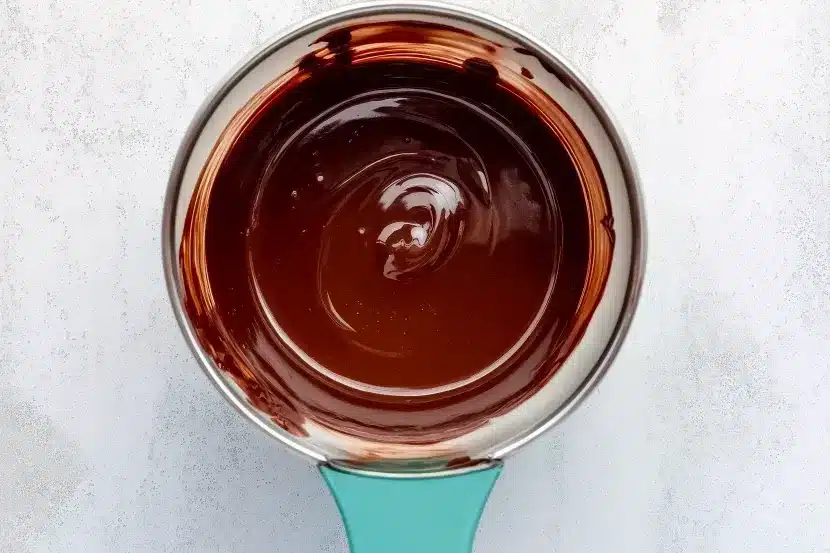 Overhead view of glass bowl with turquoise handle containing perfectly melted smooth glossy dark chocolate with silky swirl pattern and chocolate residue on bowl rim, on light marble background