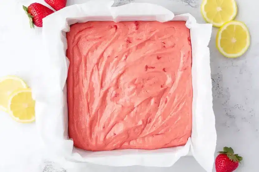 Square white parchment-lined baking pan filled with swirled coral pink strawberry brownie batter, surrounded by lemon slices and fresh strawberries on marble