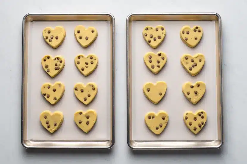 Overhead flat lay of heart-shaped chocolate chip sugar cookies on white parchment paper, mix of plain and chocolate drizzle decorated cookies, mini chocolate chips and chocolate chip pieces scattered around, wire cooling rack partially visible