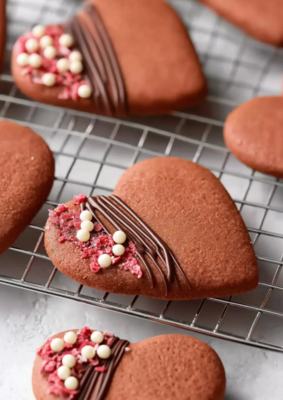 Assortment of heart-shaped chocolate Valentine cookies on wire cooling rack, some decorated with diagonal dark chocolate drizzle lines, freeze-dried raspberries and white chocolate pearls, others plain