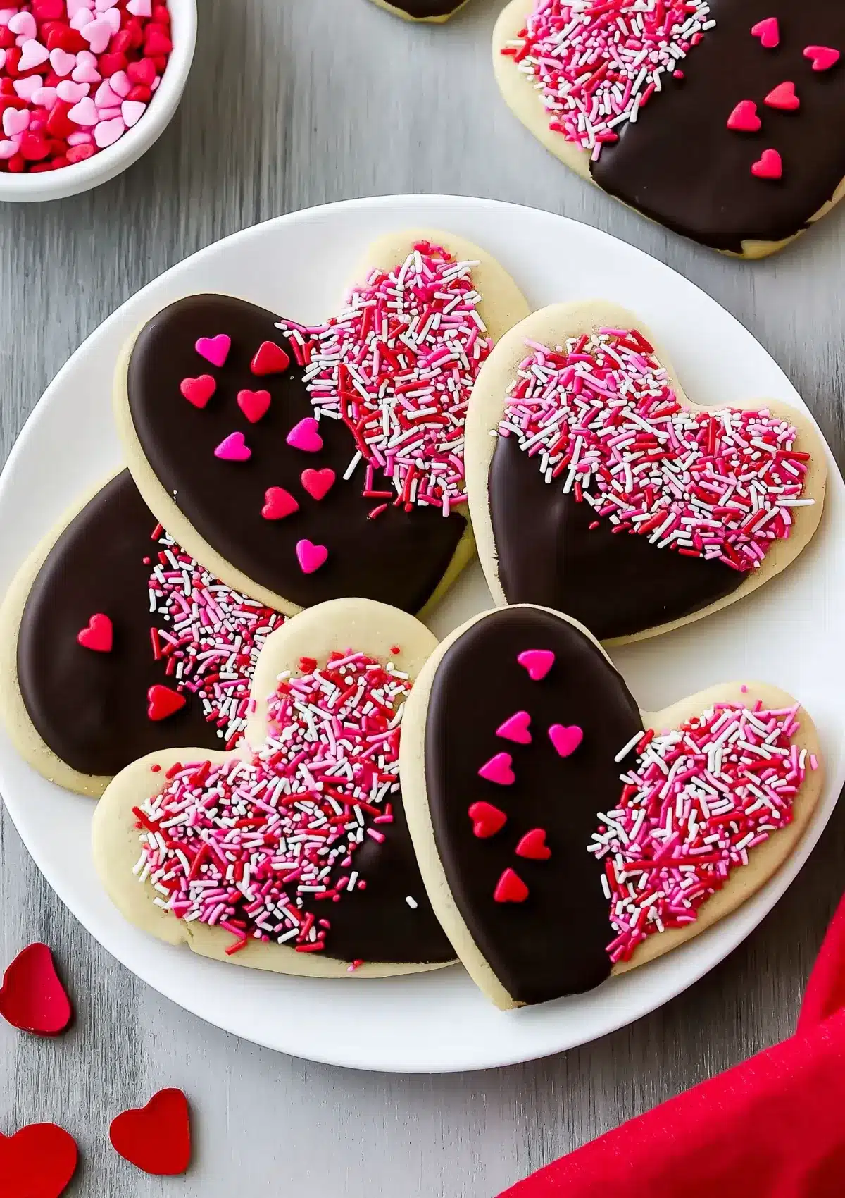 Overhead view of heart-shaped cookies with various decorations including rod sprinkles and heart-shaped candies on chocolate coating, white plate on gray wood surface