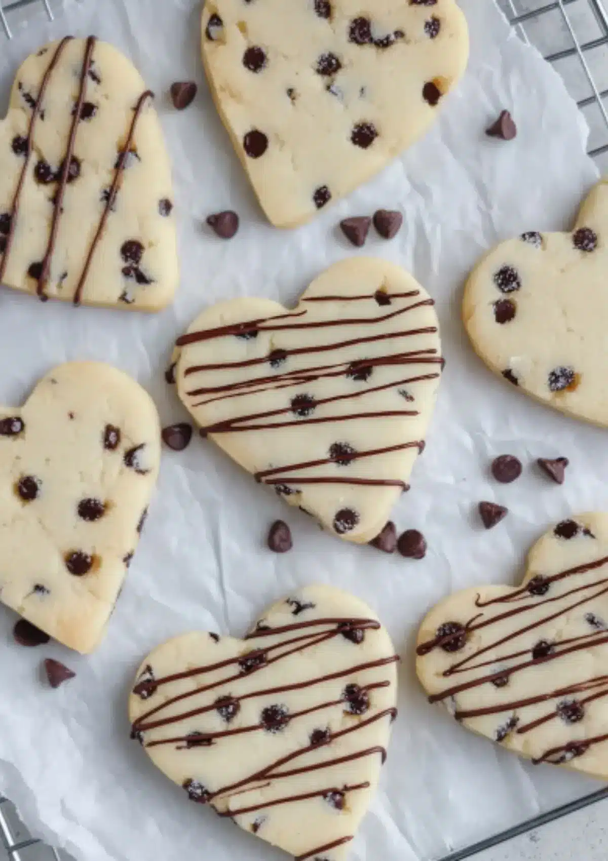 Female hand in white cable knit sweater holding heart-shaped chocolate chip sugar cookie over wire cooling rack filled with more cookies, white ceramic bowl with chocolate chips and cream linen cloth in background