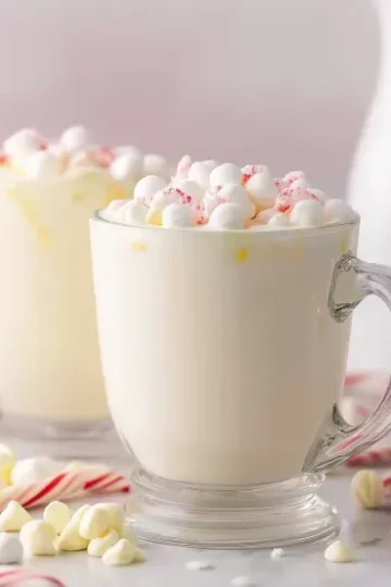 Two clear glass mugs filled with creamy white hot chocolate topped with mini marshmallows and crushed candy cane pieces, surrounded by red and white striped candy canes, red satin ribbon, white chocolate chips, and white ceramic pitchers in soft bokeh background