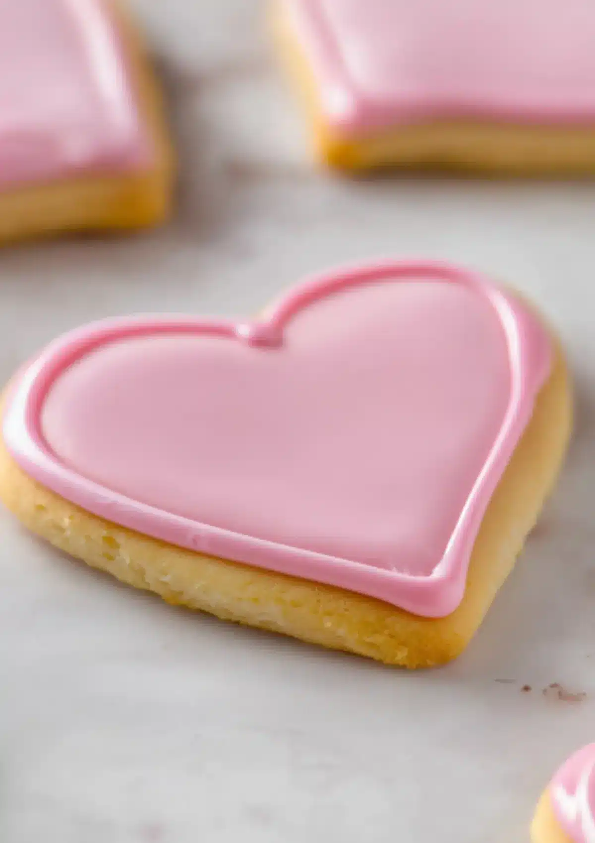 Single heart-shaped sugar cookie with pink royal icing border outline showing piping technique with soft focus cookies in background on marble countertop