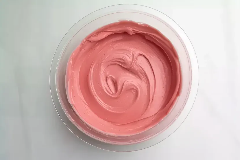 Overhead shot of smooth coral pink royal icing in clear glass bowl showing swirled mixing texture on white surface