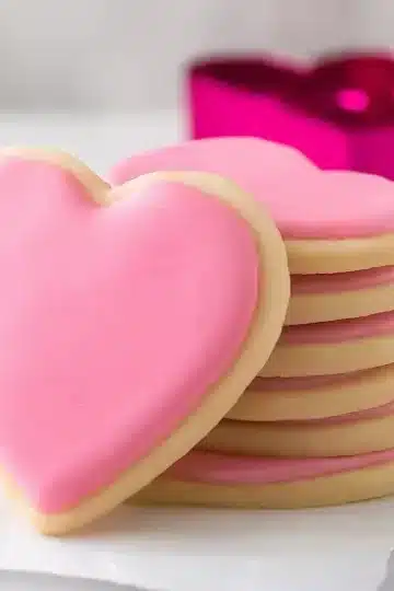 Stack of heart-shaped sugar cookies with pink royal icing on white marble surface with bright magenta heart-shaped cookie cutter in soft focus background