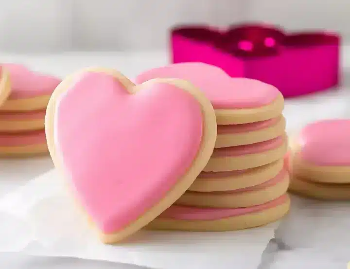 Stack of heart-shaped sugar cookies with pink royal icing on white marble surface with bright magenta heart-shaped cookie cutter in soft focus background